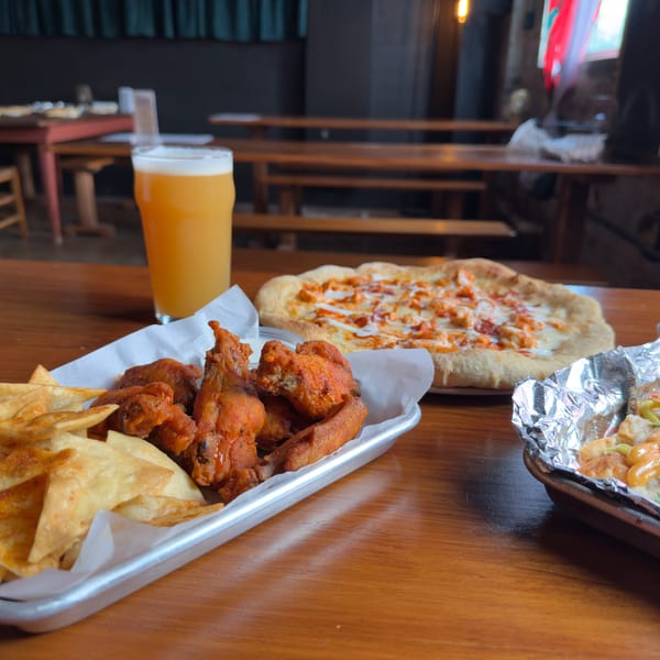A table with a beer, buffalo chicken pizza, wings, chips, and loaded fries.