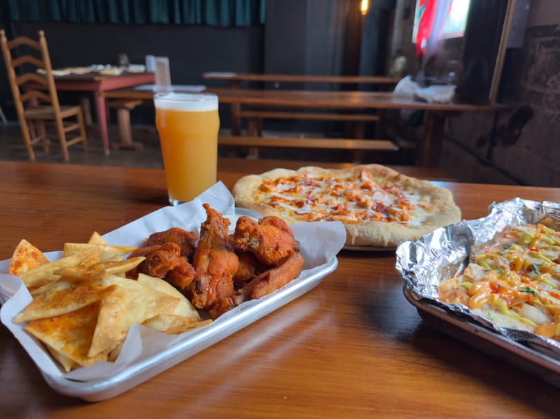 A table with a beer, buffalo chicken pizza, wings, chips, and loaded fries.