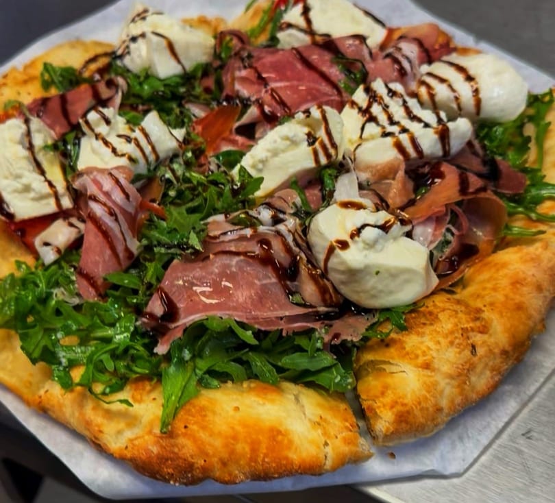 A gourmet pizza topped with prosciutto, fresh mozzarella, arugula, and balsamic glaze.