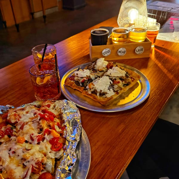 Two pizzas, two cocktails, and a beer flight on a wooden bar with a glowing lamp.