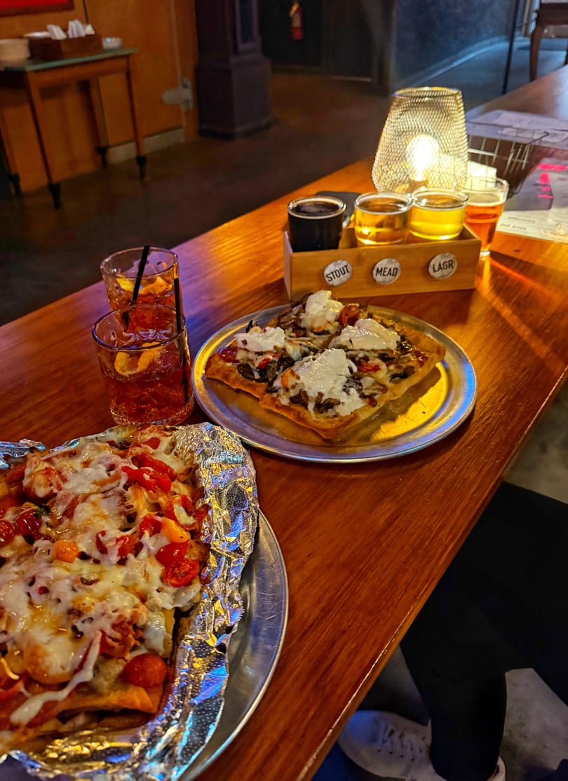 Two pizzas, two cocktails, and a beer flight on a wooden bar with a glowing lamp.